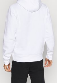 Biała hoodie o miękkiej teksturze, z kapturem na sznurku, ściśniętymi mankietami i luźnym krojem. Tył jest gładki, z widocznymi detalami szycia.