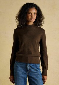 Brauner Pullover mit langen Ärmeln, ribbeliger Textur und hohem Ausschnitt; verfügt über Knöpfe an den Ärmeln und hat eine lockere Passform. Kombiniert mit blauen Jeans.