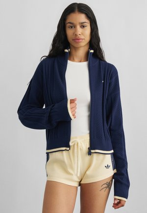 Junge Frau mit langen dunklen Haaren, die einen marineblauen Reißverschluss-Pullover, ein weißes Oberteil und beige Adidas-Shorts trägt, mit einem Spinnen-Tattoo am Oberschenkel.