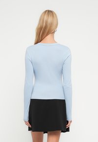 Γυναίκα με μακριά ξανθά μαλλιά που φοράει ανοιχτό μπλε μακρυμάνικη μπλούζα και μαύρη φούστα, στέκεται με την πλάτη προς το μέρος μας μπροστά σε λευκό φόντο.