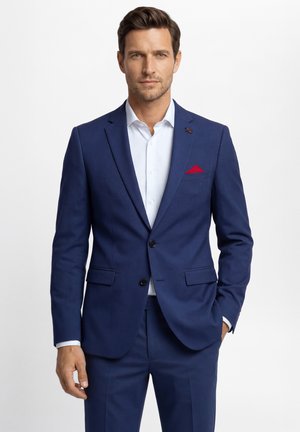 Man met bruin haar draagt een blauw pak met een wit overhemd en een rood pochet, staand tegen een effen witte achtergrond.
