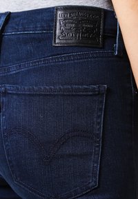 Jeans de mezclilla azul oscuro con una etiqueta de cuero negro que presenta un logo y texto en relieve. Costura notable y estilo de bolsillos en la parte trasera.