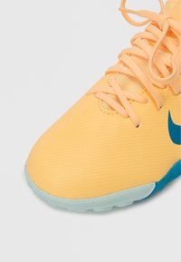 Zapatilla deportiva amarilla con tejido texturizado, puntera redondeada y acentos en turquesa. Presenta cordones planos y una suela azul claro.