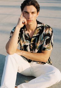 Young man with dark hair wearing a black tropical print shirt and white pants sits cross-legged outdoors, resting head on hand, looking at camera.