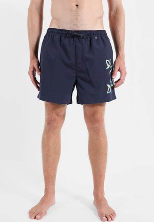 Homme portant un short de bain bleu marine avec un motif floral multicolore sur la cuisse droite, debout pieds nus devant un fond uni.