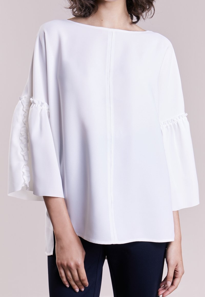 Femme portant un chemisier blanc ample à larges manches avec des détails de volants froncés, associée à un pantalon foncé, debout devant un fond uni.