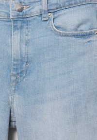 Blugi din denim albastru deschis, prim-plan, arătând buzunarul frontal, bucla pentru curea, cusătura și nasturele metalic cu gravura brandului.