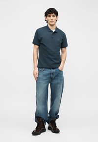 Mladý muž stojaci s jednou rukou v vrecku, oblečený v tmavomodrej polokošeli, voľných vyblednutých džínsach vyhrnutých na koncoch a tmavohnedých šnurovacích topánkach.