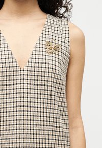 Γυναίκα που φοράει αμάνικο μπεζ και μαύρο καρό φόρεμα με μια χρυσή καρφίτσα σε σχήμα λουλουδιού καρφωμένη κοντά στον ώμο.