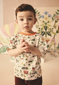 Børne t-shirt med korte ærmer, cremefarvet med farverige dyre- og naturmønstre, har en orange krave og løs pasform. Brune cordbukser.