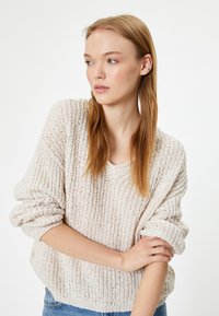 Maglione lavorato a maglia di un beige chiaro con una vestibilità ampia e morbida, texture a costine e polsini arrotolati. Indossato con jeans di denim blu.