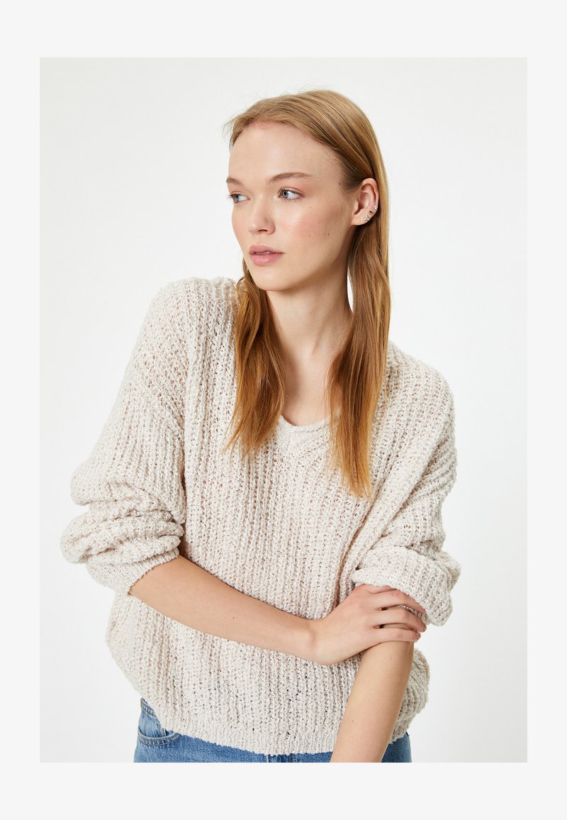 Giovane donna con lunghi capelli rossi che indossa un maglione crema strutturato, braccia incrociate, che guarda verso sinistra contro uno sfondo bianco uniforme.