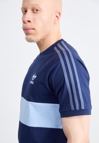 Mornarica plava majica s lagano plavom horizontalnom prugom. Ima tri lagane plave pruge na ramenima i Adidas logo na prsima.