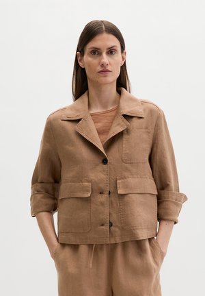 Frau trägt eine hellbraune, geknöpfte Jacke mit hochgekrempelten Ärmeln, passende Hose und ein braunes Hemd, steht vor einem schlichten Hintergrund.