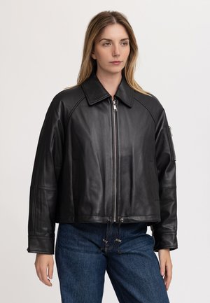Frau trägt eine schwarze Lederjacke mit Reißverschluss und Kragen, kombiniert mit blauen Jeans, steht vor einem schlichten hellen Hintergrund.