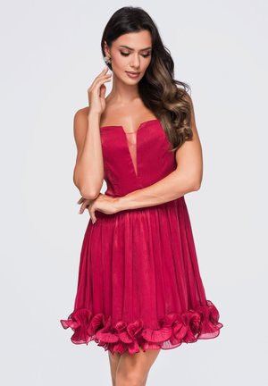 Femme aux longs cheveux ondulés portant une robe rouge sans manches avec un décolleté en V profond et un ourlet volanté, posant avec une main près du visage.