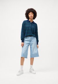 Pulover s kratkimi rokavi v mornarsko modrih črtah in ovratnikom, v kombinaciji s svetloblujimi denim kratkimi hlačami. Bele teniske in nogavice dopolnjujejo videz.