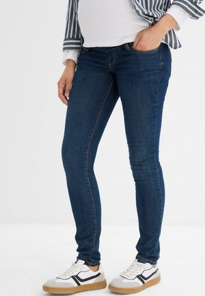 Jean skinny bleu foncé avec une texture denim lisse, comportant cinq poches et des coutures subtiles, associés à des baskets blanches.
