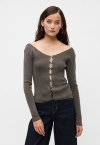 Νέα γυναίκα με μακριά σκούρα μαλλιά φοράει γκρίζα ριγωτή κάρντιγκαν με κουμπιά και σκούρα μπλε τζιν, ποζάροντας μπροστά σε λευκό φόντο.