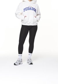Felpa bianca con testo "PEGADOR" blu, leggings neri, calze bianche e sneakers grigie. La felpa presenta una tasca a marsupio e una vestibilità rilassata.