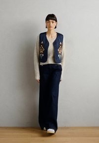 Džinsu veste ar krāsainiem ziedu izšuvumiem virs gaiša, struktūrēta džemperi. Apvienota ar tumši zilām plašām biksēm un baltām kurpēm.