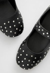 Ballerines Mary Jane en cuir noir avec des rivets en argent sur l'avant. Forme de bout arrondi avec une bride sur le cou-de-pied. Texture lisse.