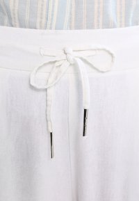 Pantalons en lin blanc avec taille à cordon. Le cordon est doté d'aglets en métal argenté. Le tissu a une texture légère et est respirant.