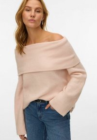 Frau in einem hellpinken, schulterfreien Pullover mit weitem Schnitt und blauen Jeans, die mit einer Hand in der Tasche vor einem weißen Hintergrund posiert.