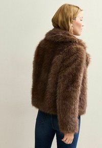 Veste cropped en fausse fourrure marron avec une texture douce, un large col et des manches longues. Portée sur un jean bleu foncé, vue de dos.