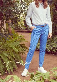 Pantalons taillés bleus avec une texture lisse, associés à une chemise à rayures et des baskets blanches, le tout sur fond de verdure luxuriante.