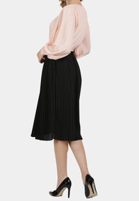 Vrouw in een lichtroze geplooide blouse met lange mouwen, zwarte geplooide midirokken en zwarte hoge hakken staat zijwaarts.