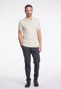 Beige kortärmad polo med krage, i kombination med mörkgrå slim-fit byxor och svarta läderskor mot en enkel bakgrund.
