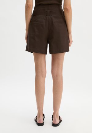 Vrouw draagt een donkere bruine shorts met hoge taille en achterzakken, gecombineerd met zwarte sandalen, staand tegen een effen witte achtergrond.