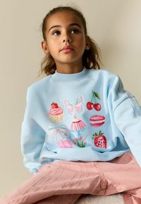 Sweat-shirt bleu clair avec des desserts brodés colorés et un papillon ; associé à un pantalon rose texturé. Tissu lisse, col rond.