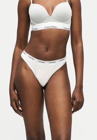 Γυναίκα που φοράει ανοιχτό γκρι και λευκό σουτιέν και εσώρουχο Calvin Klein με ελαστικές ταινίες με την επωνυμία, στέκεται μπροστά σε απλό λευκό φόντο.