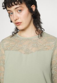 Blusa verde-claro com um design de renda nos ombros e no peito, feita de um tecido suave e texturizado abaixo da renda.