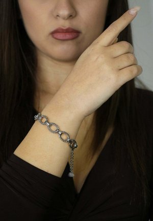 Bracelet en métal argenté avec des maillons circulaires entrelacés, une extension de chaîne et un pendentif, porté sur un poignet à peau claire.