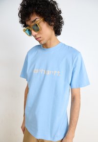 Light blue cotton t-shirt features a beige logo on the front. The model also wears oversized yellow sunglasses with tinted lenses.