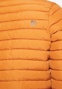 Veste matelassée orange avec coutures horizontales et un petit logo géométrique gris sur la zone de la poitrine.