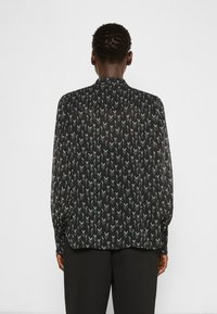 Zwarte blouse met lange mouwen en een delicaat patroon van kleine, puntige vormen in gedempte kleuren. Lichtgewicht, doorzichtige stof met een hoge halsontwerp.