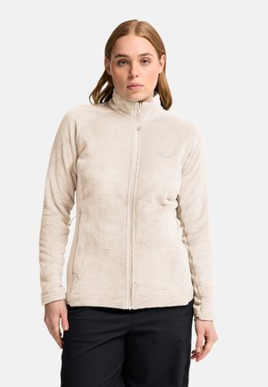 Frau trägt eine beige Fleecejacke mit durchgehendem Reißverschluss und schwarze Hose, blickt nach vorne vor einem schlichten weißen Hintergrund.