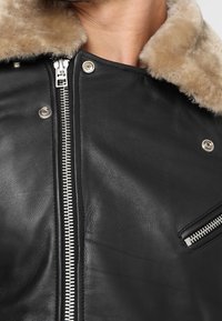 Giacca di pelle nera con zip e bottoni argentati, caratterizzata da un colletto in shearling beige e una tasca sul petto con zip.