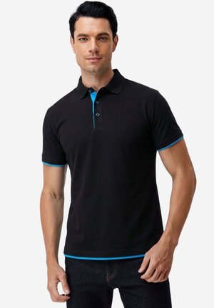 Mann trägt schwarzes Poloshirt mit blauem Besatz an Kragen, Ärmeln und Saum, steht vor weißem Hintergrund.