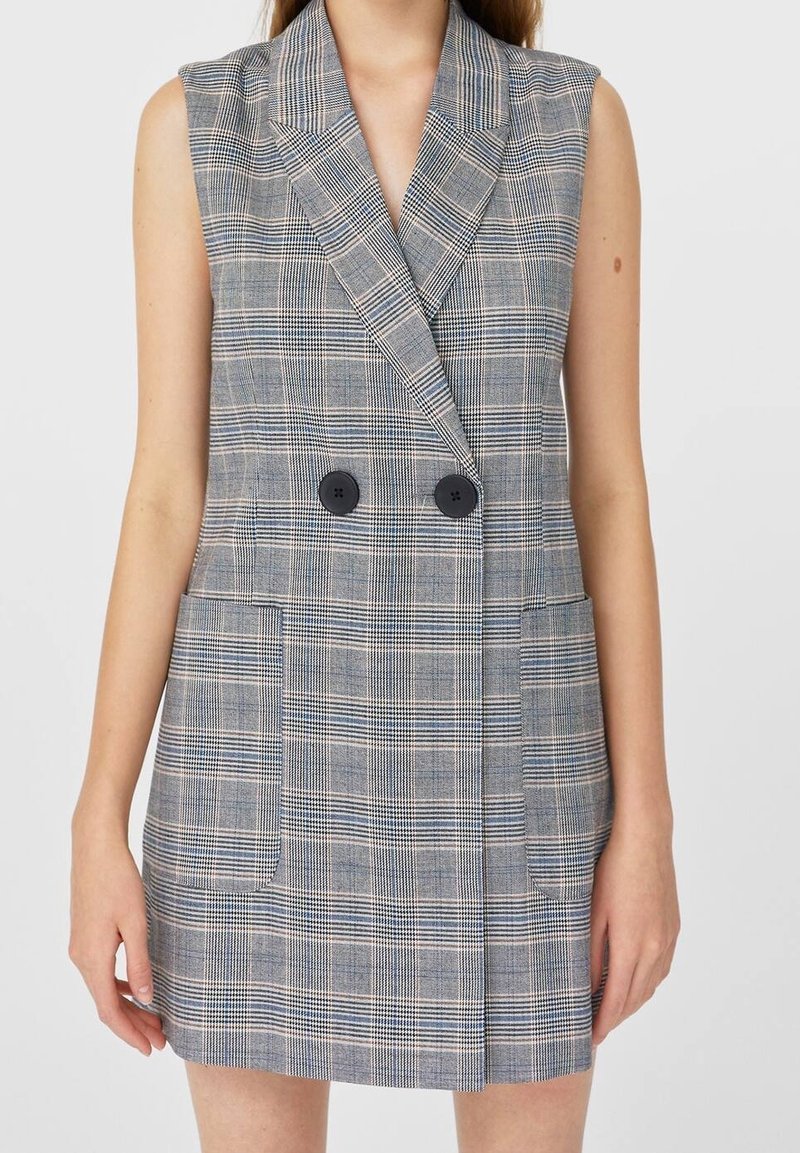 Vestido blazer sin mangas de doble botonadura en un patrón de cuadros grises, con grandes botones negros y dos bolsillos delanteros.