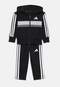Crni dukserica i trenirke set s bijelim prugama i Adidas logotipom, dizajniran za aktivnu odjeću, prikazan na bijeloj pozadini.