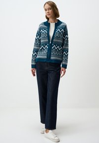 Gilet zippé teal avec un motif fair isle en blanc et en bleu foncé, col et poignets côtelés. Assorti à un jean bleu foncé à jambes larges et des baskets blanches.