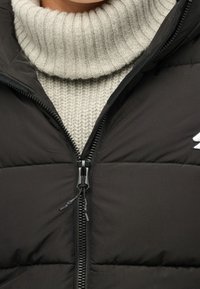 Musta toppaliivi korkealla kaulaosalla ja sen alla teksturoidulla harmaalla poolopaidalla. Liivissä on etuvetoketju ja pieni brändäysyksityiskohta.