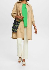 Un trench coat beige sopra un top verde brillante, abbinato a pantaloni a zampa d'elefante color crema e mules beige. L'accessorio è una borsa blu navy con motivo a griglia verde.