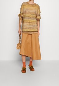 Pull en maille aux tons chauds avec une coupe ample, associé à une jupe marron longueur genou, des chaussettes oranges, et des sandales marron à brides. Petit sac à main inclus.