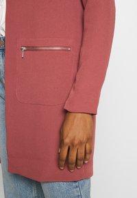 Žebrovaná růžová bunda s kapsou na zip; dlouhé rukávy; texturovaná tkanina; nošena přes světle modré džíny, ukazující detaily ruky a paže.